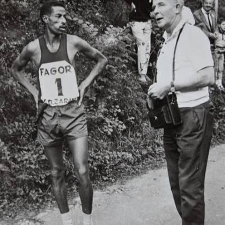 Abebe Bikila, wearing race bib number 1, stands with hands on hips beside Onni Niskanen at the roadside.
