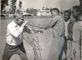 Onni Niskanen and Abebe Bikila in a playful boxing pose on a training field as athletes look on.